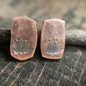 May include: A pair of copper earrings with a lotus flower design. The earrings are rectangular in shape and have a hammered texture.