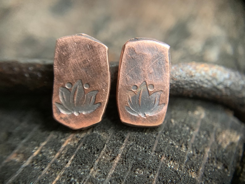 May include: A pair of copper earrings with a lotus flower design. The earrings are rectangular in shape and have a hammered texture.