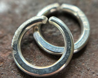 May include: Two silver jump rings, slightly open, are positioned on a textured, brown surface. The rings are circular, with a bright, reflective finish. The image is a close-up, highlighting the metal's sheen and the rings' construction.