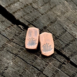 May include: A pair of copper earrings with a lotus flower design. The earrings are a rectangular shape and have a rustic, oxidized finish.