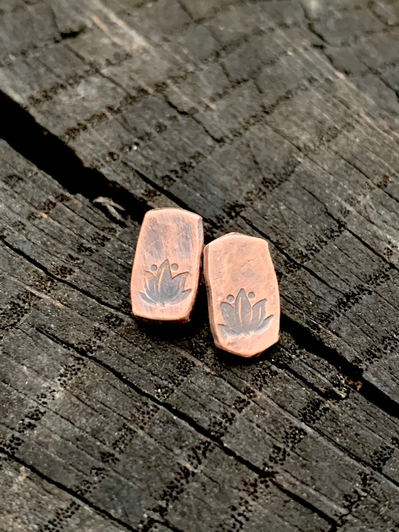 May include: A pair of copper earrings with a lotus flower design. The earrings are a rectangular shape and have a rustic, oxidized finish.