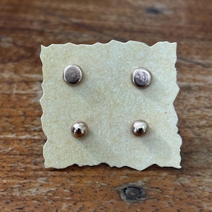 May include: Two pairs of small, round, metallic stud earrings with a polished rose gold finish. The four individual earrings are arranged in a square formation, secured onto a square piece of light beige paper with deckled, uneven edges.