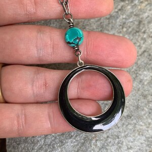 May include: Silver-toned necklace featuring a turquoise bead and a large, black enamel circle pendant. The chain is a textured link style. The necklace is held against a gray stone surface.