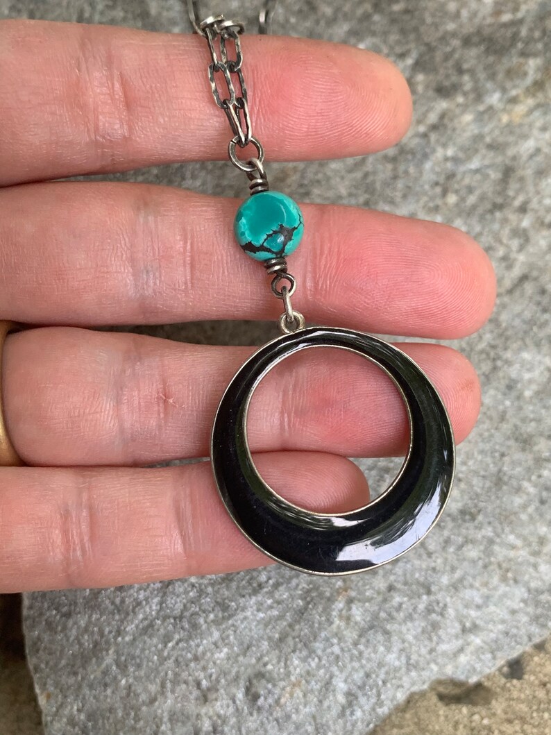 May include: Silver-toned necklace featuring a turquoise bead and a large, black enamel circle pendant. The chain is a textured link style. The necklace is held against a gray stone surface.