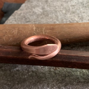 Puede incluir: Un anillo de cobre con forma de serpiente, con un acabado liso y pulido. El anillo est&aacute; apoyado sobre una superficie de madera.