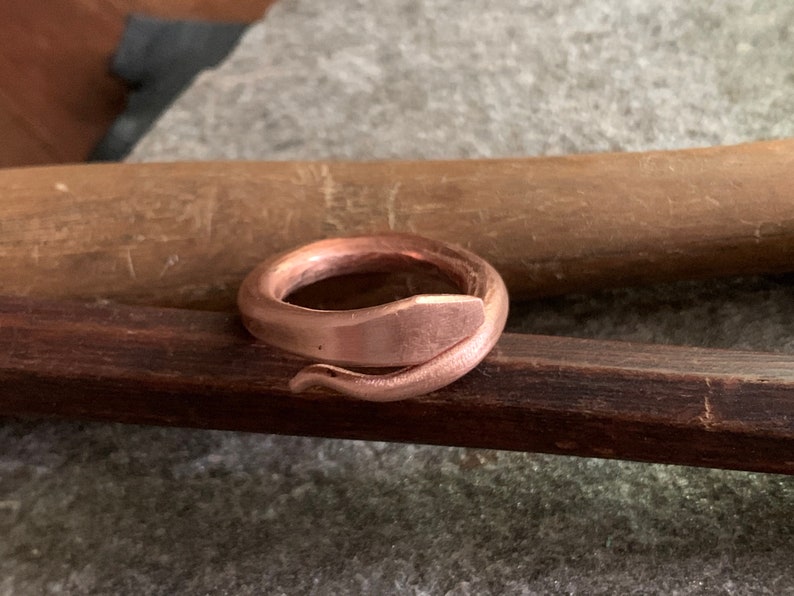 Puede incluir: Un anillo de cobre con forma de serpiente, con un acabado liso y pulido. El anillo est&aacute; apoyado sobre una superficie de madera.