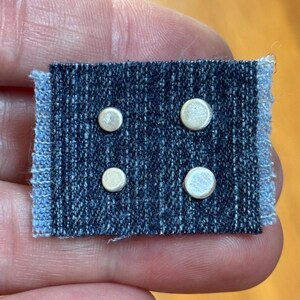 May include: A small rectangular piece of dark blue denim fabric with frayed edges on the left and right sides. Four circular, flat, silver-colored metal studs are arranged in a two-by-two grid pattern on the surface of the denim material.