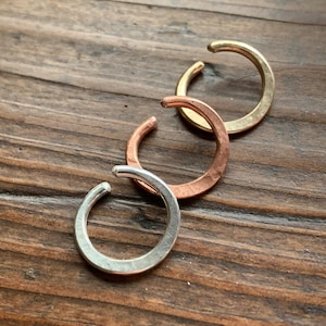 May include: Three ear cuffs in gold, copper, and silver tones are displayed on a wooden surface. The open-ended, circular cuffs are designed to clip onto the ear. The gold cuff is at the top, followed by copper and silver.