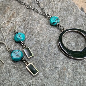 May include: A silver necklace and matching earrings set. The necklace features a large, dark ring pendant and turquoise beads. The earrings have turquoise beads and rectangular black accents. The jewelry is displayed on a gray stone surface.
