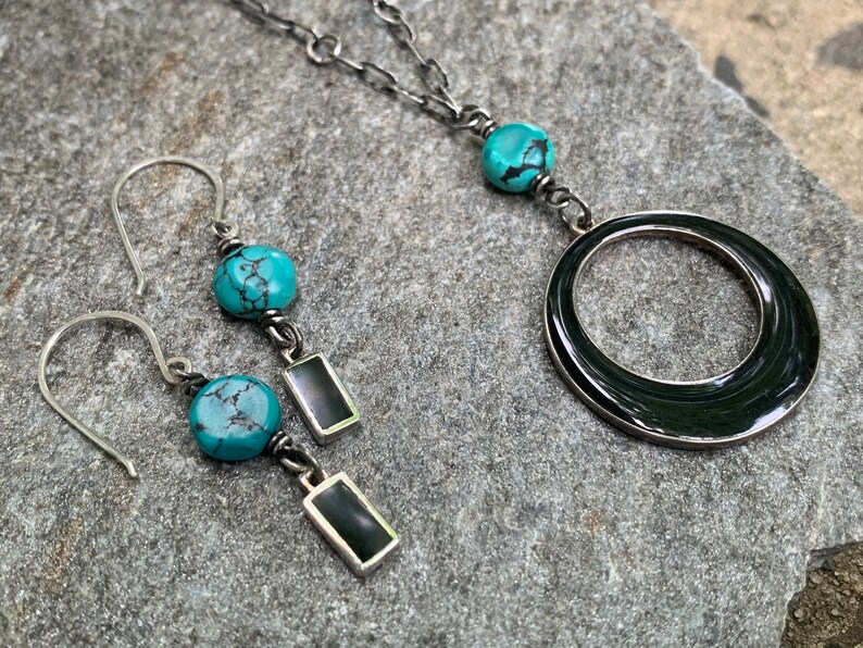 May include: A silver necklace and matching earrings set. The necklace features a large, dark ring pendant and turquoise beads. The earrings have turquoise beads and rectangular black accents. The jewelry is displayed on a gray stone surface.