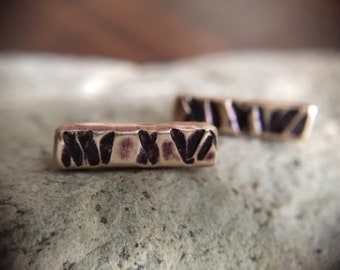 May include: Two small, gold-toned bar earrings with a textured, geometric design. The earrings are made of metal and have a dark brown finish.