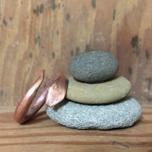 Puede incluir: Un anillo de cobre con un dise&ntilde;o &uacute;nico y angular. El anillo est&aacute; apoyado en una pila de tres piedras grises y marrones.