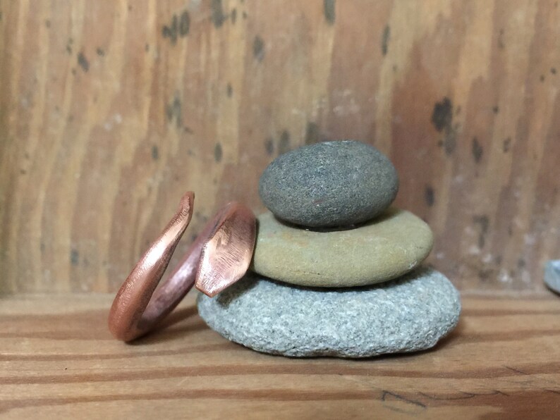 Puede incluir: Un anillo de cobre con un dise&ntilde;o &uacute;nico y angular. El anillo est&aacute; apoyado en una pila de tres piedras grises y marrones.