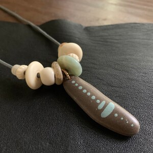 May include: A gray cord necklace with white and light green beads and a brown stone pendant with white painted dots and lines.