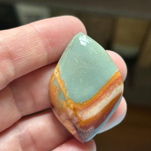 May include: A teardrop-shaped gemstone with a polished surface. The stone features a blend of colors, including light blue, orange, red, and white, creating a layered effect. The gemstone is held in a hand.