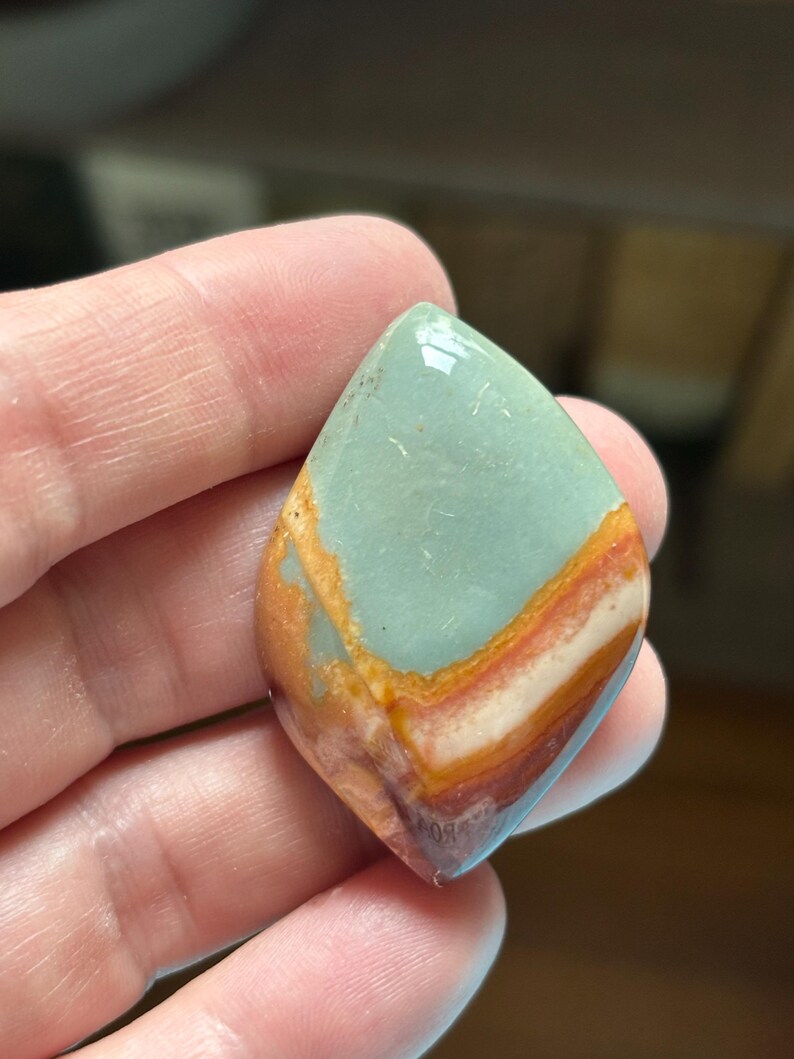 May include: A teardrop-shaped gemstone with a polished surface. The stone features a blend of colors, including light blue, orange, red, and white, creating a layered effect. The gemstone is held in a hand.