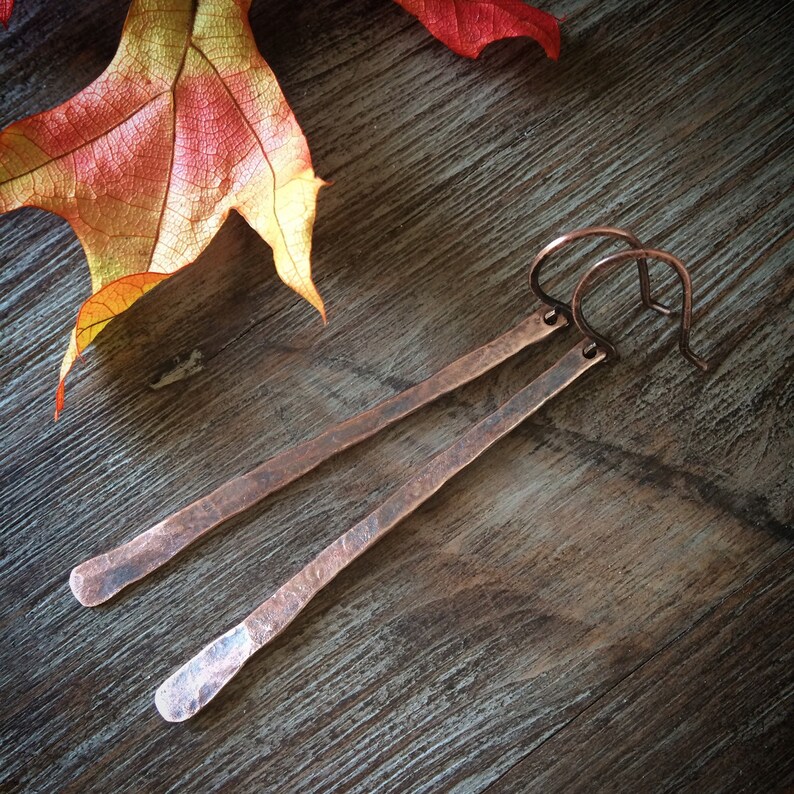 May include: A pair of long, hammered copper earrings with a simple, minimalist design. The earrings are a warm, reddish-brown color and have a slightly textured finish.