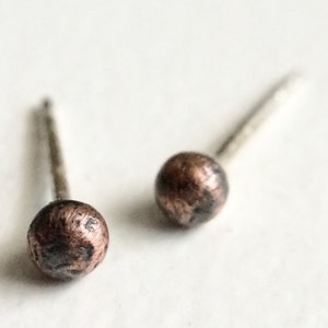 May include: A pair of stud earrings featuring small, spherical, textured copper-colored beads attached to thin, straight metal posts. The surface of each round bead displays a mottled, dark and light brown finish with a slightly uneven, rustic appearance.
