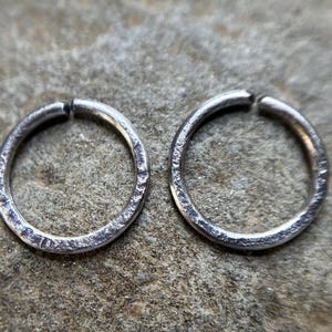 May include: Two silver-colored, hammered-texture hoop earrings. The earrings are circular with a small gap at the top. The earrings are displayed on a rough, gray stone surface.