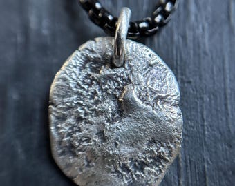 May include: A silver pendant with a textured, irregular surface hangs from a black chain. The pendant is oval-shaped and has a loop at the top for the chain. The background is a dark, textured surface.