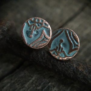 May include: A pair of round, copper earrings with a teal blue enamel finish. The earrings feature a unique, abstract design.
