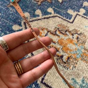 May include: A long, thin, copper hair stick with a turquoise stone at the top. The stick is slightly curved and has a textured surface.