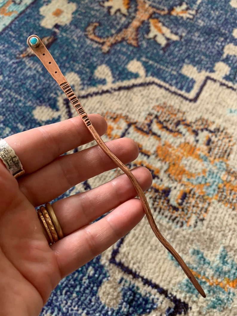 May include: A long, thin, copper hair stick with a turquoise stone at the top. The stick is slightly curved and has a textured surface.