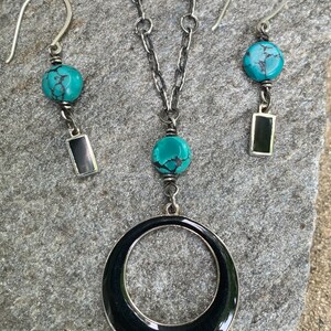 May include: A silver necklace and earring set featuring turquoise beads and black rectangular accents. The necklace has a large black circular pendant. The earrings and necklace have a matching design.