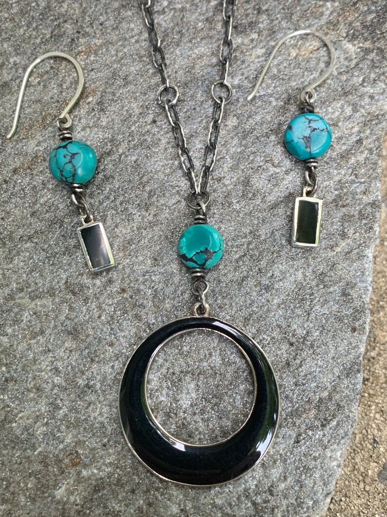 May include: A silver necklace and earring set featuring turquoise beads and black rectangular accents. The necklace has a large black circular pendant. The earrings and necklace have a matching design.