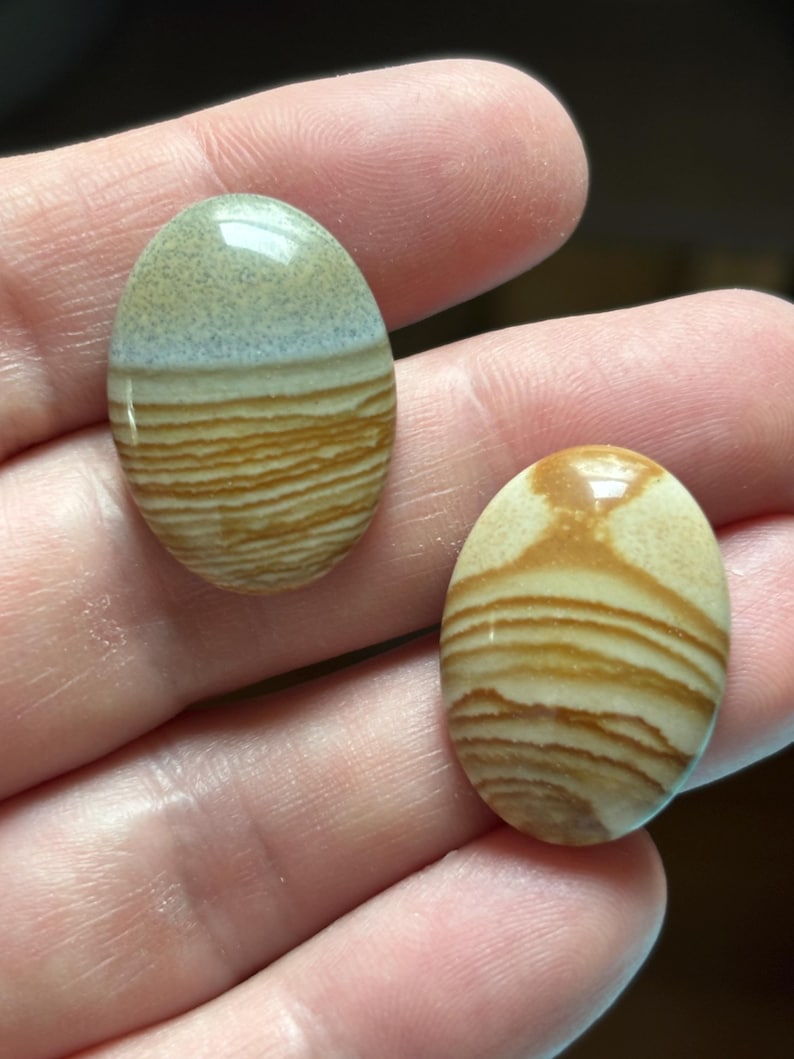 Puede incluir: Dos piedras preciosas ovaladas con un patr&oacute;n en capas y bandas en tonos beige, marr&oacute;n y crema. Las piedras se sostienen en una mano, mostrando sus superficies pulidas y variaciones naturales.