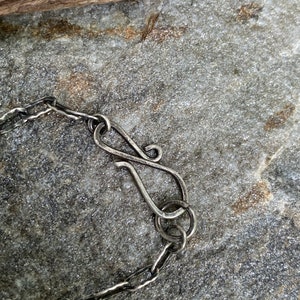 May include: Close-up of a silver necklace clasp and chain. The clasp is a stylized S-hook, connected to a chain with textured links. The jewelry rests on a gray stone surface, with a piece of wood in the background.