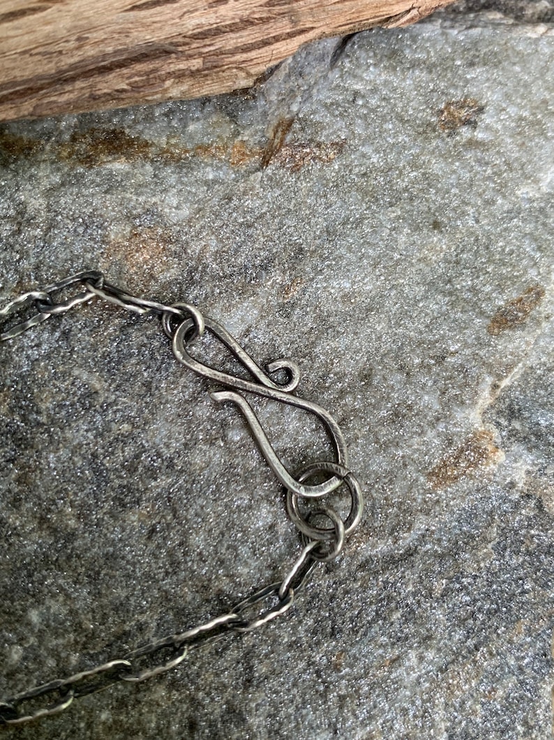 May include: Close-up of a silver necklace clasp and chain. The clasp is a stylized S-hook, connected to a chain with textured links. The jewelry rests on a gray stone surface, with a piece of wood in the background.