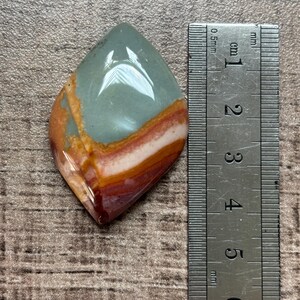 May include: A polished gemstone with a teardrop shape. The stone features bands of color, including teal, orange, white, and maroon. A ruler is placed next to the stone, showing measurements in centimeters.