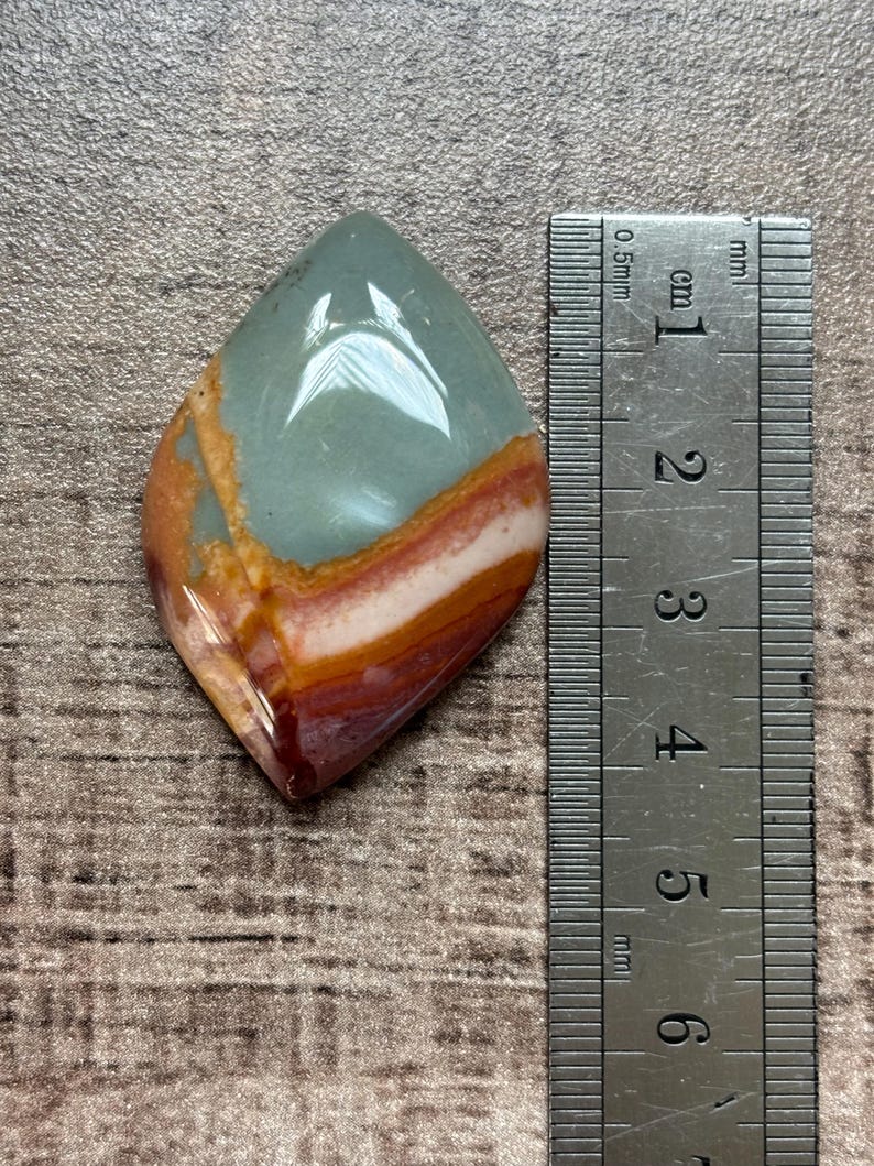 May include: A polished gemstone with a teardrop shape. The stone features bands of color, including teal, orange, white, and maroon. A ruler is placed next to the stone, showing measurements in centimeters.