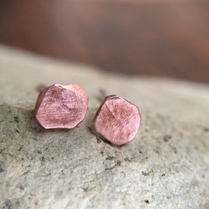 May include: A pair of rose gold stud earrings with a geometric, faceted design. The earrings are made of metal and have a brushed finish.
