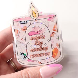 May include: A clear sticker in the shape of a candle with a pink and white design. The sticker has the text "smells like cowboy romance" and a pink cowboy hat graphic. The sticker is held by a hand with a pink and white manicure.