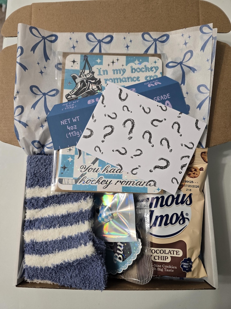 May include: An open cardboard box filled with various items. Includes a blue and white striped sock, a card with a hockey skate graphic, and a card with question marks. Also contains a box with "NET WT 4oz (113g)" printed on it, and a package of chocolate chip cookies.
