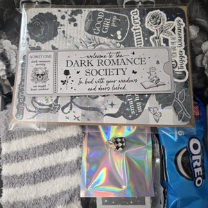 May include: A gift set featuring a variety of items. Includes a sticker sheet with gothic-themed designs, a pair of gray and white striped socks, a small iridescent package with a checkered charm, and a package of Oreo cookies.
