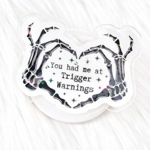 May include: A clear, heart-shaped sticker featuring black skeleton hands forming a heart shape. The sticker has the text "You had me at Trigger Warnings" with star and glitter accents. The sticker is on a white background.