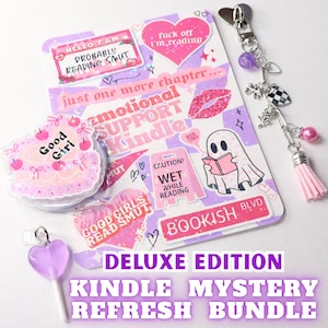 May include: A Deluxe Edition Kindle Mystery Refresh Bundle featuring stickers, a keychain, and a lollipop. The stickers have phrases like "Emotional Support Kindle" and "Good Girls Read Smut." The keychain has charms and a pink tassel. The lollipop is heart-shaped.