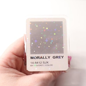 May include: A clear, rectangular sticker with the text "MORALLY GREY" and "MY FAVORITE COLOR". The sticker has a gray background with colorful star-shaped glitter. The sticker's color code is 14-8412 SJX.