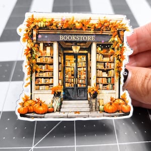 May include: A die-cut acrylic sticker featuring a bookstore storefront decorated with orange autumn leaves and pumpkins. The shop has glass doors, shelves filled with books, and a sign that reads BOOKSTORE. The design includes holographic accents.