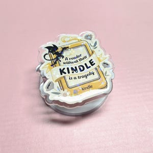 May include: A clear, circular phone grip with a decorative design. The design features a yellow Kindle with the text "A reader without their Kindle is a tragedy." A black dragon and floral accents complete the design.
