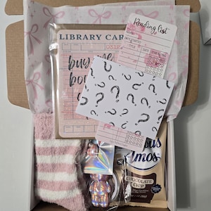 May include: A gift box filled with pink and white striped socks, a library card, a reading list card, a card with question marks, a holographic bear keychain, and a chocolate chip cookie. The box is lined with pink bow-patterned tissue paper.