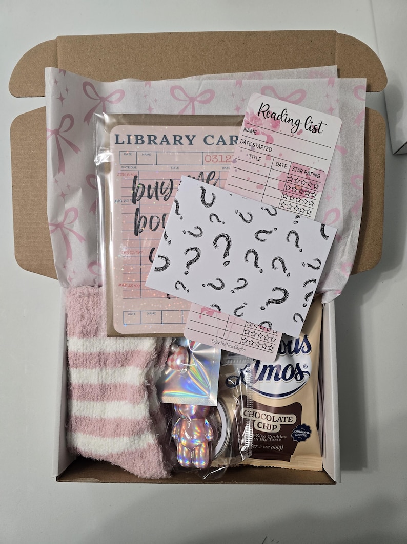 May include: A gift box filled with pink and white striped socks, a library card, a reading list card, a card with question marks, a holographic bear keychain, and a chocolate chip cookie. The box is lined with pink bow-patterned tissue paper.