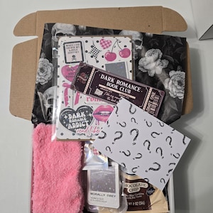 May include: An open cardboard box filled with themed items. Includes a pink fuzzy sock, a notebook with cherries, a ticket that says "Dark Romance Book Club", a packet of "Morally Grey" color, and a chocolate chip cookie.