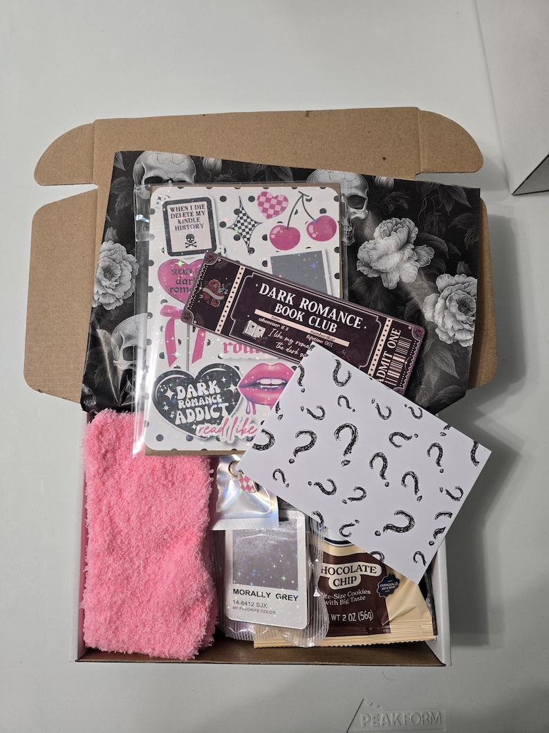 May include: An open cardboard box filled with themed items. Includes a pink fuzzy sock, a notebook with cherries, a ticket that says "Dark Romance Book Club", a packet of "Morally Grey" color, and a chocolate chip cookie.