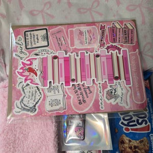 May include: A pink and white sticker sheet with book-themed designs, including text like "I love books" and "Trigger Warnings." The sheet is in a clear plastic sleeve, with a pink fluffy item and a bag of cookies visible.