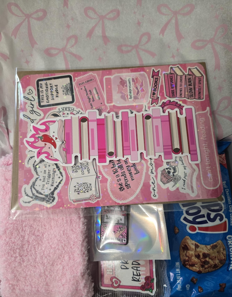 May include: A pink and white sticker sheet with book-themed designs, including text like "I love books" and "Trigger Warnings." The sheet is in a clear plastic sleeve, with a pink fluffy item and a bag of cookies visible.