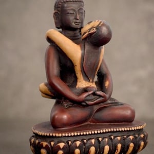 Può includere: Statua in legno dettagliata di Buddha in posizione meditativa seduta, che abbraccia un'altra figura. La statua è principalmente marrone scuro con accenti beige chiaro e poggia su una base decorativa. Il tema è spirituale e artisto.